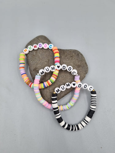 Hier sieht man handgemachte Armbänder in den Farben Bunt, Pastell und Schwarz-Weiß aus Perlenscheiben. Mum Doc Perlenbuchstaben sind in der Mitte aufgereiht.